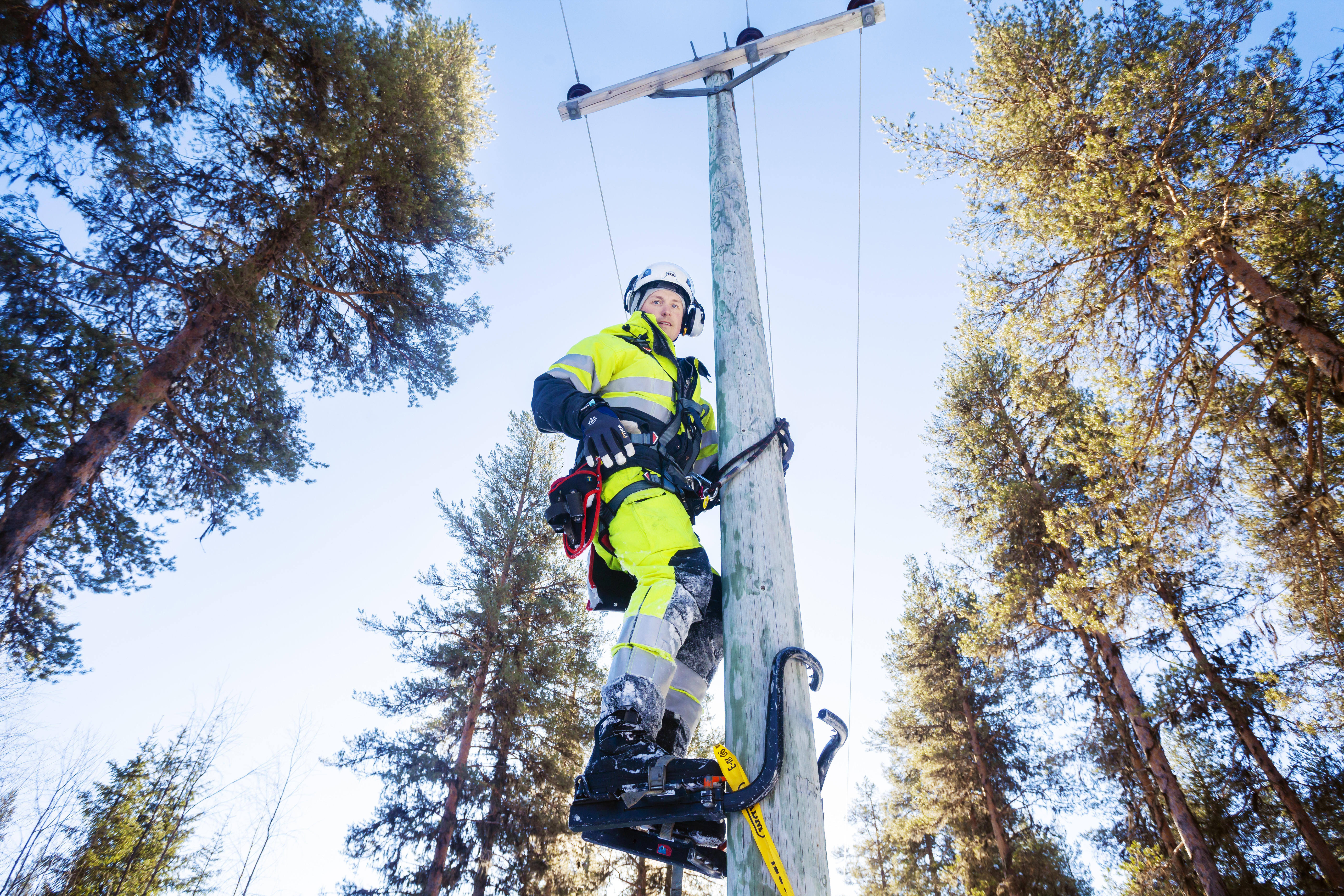 En arbetare i varselkläder, hjälm och stegjärn i elstolpe