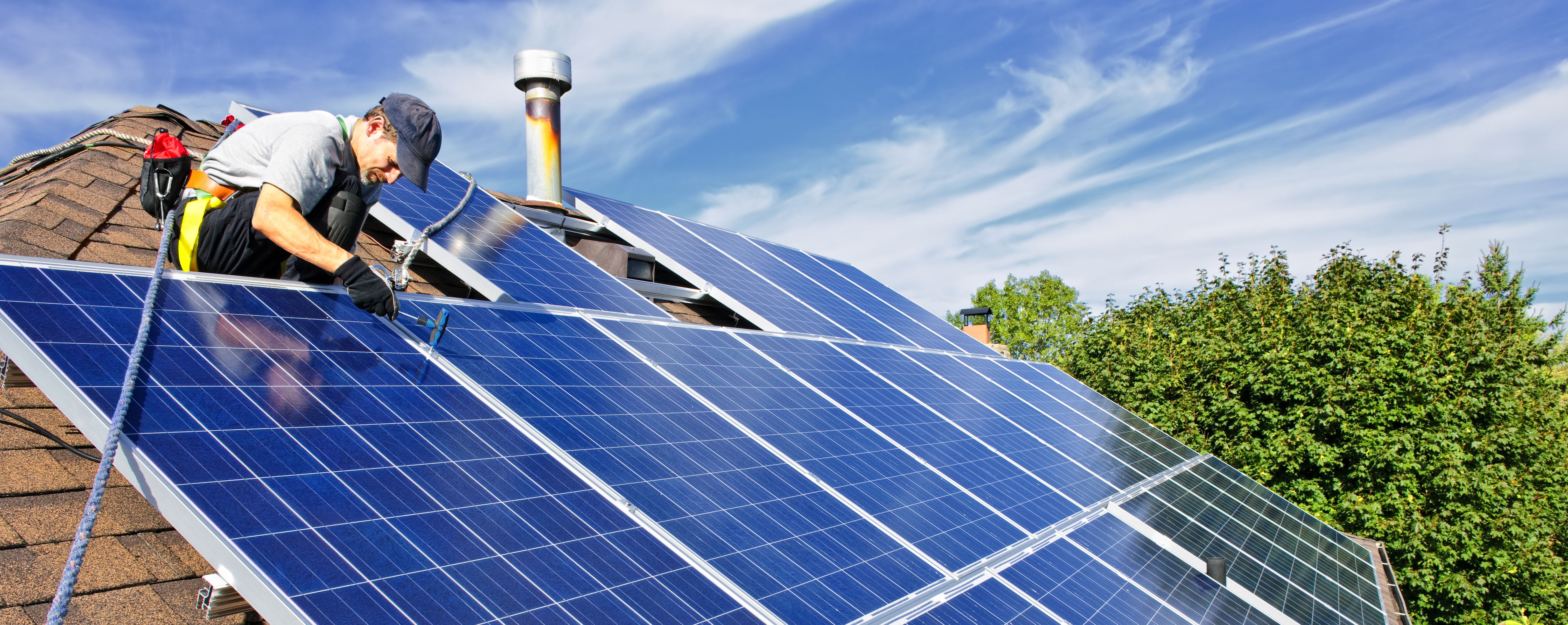 Man i keps sitter fast i en sele på ett tak med solpaneler. Foto