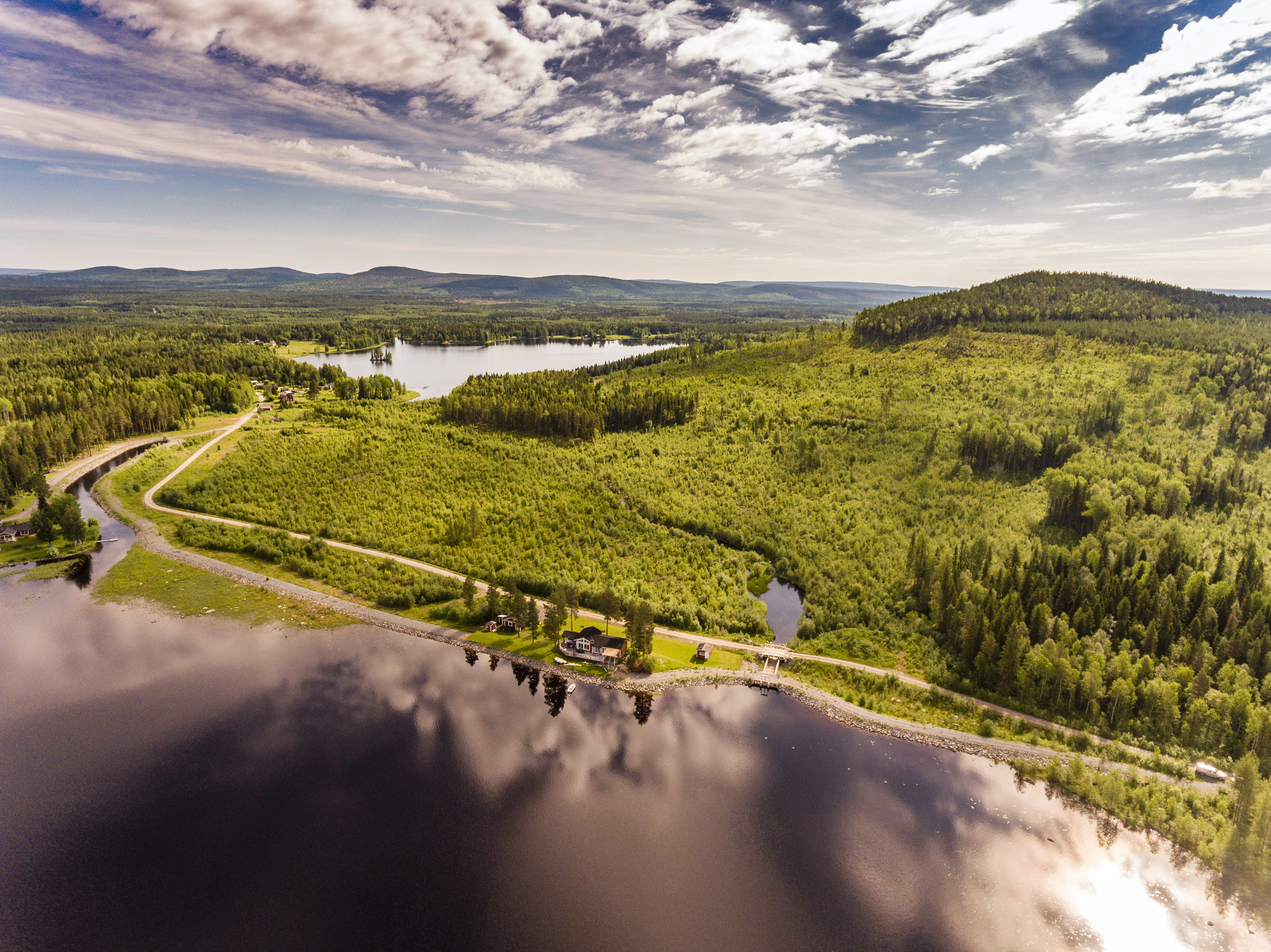 Blå himmel med moln, berg i bakgrunden och skog, väg och vatten. Foto