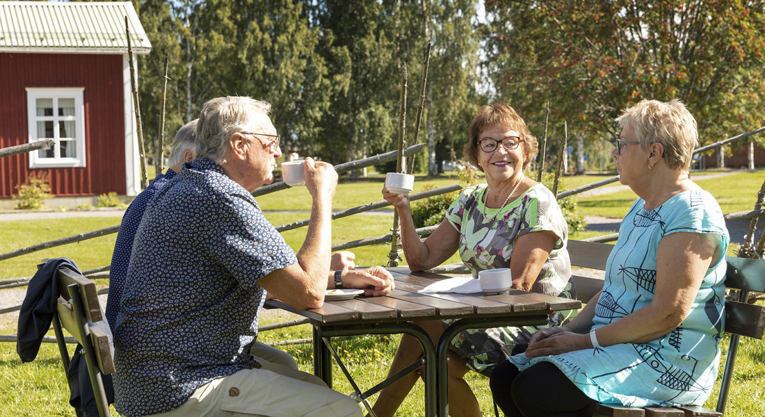 Fyra äldre personer dricker kaffe i trädgården. Rött hus i bakgrunden