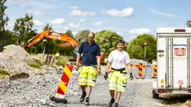 Man och kvinna i arbetskläder går på grus mellan vägskyltar och en bil. I bakgrunden syns en grävmaskin.