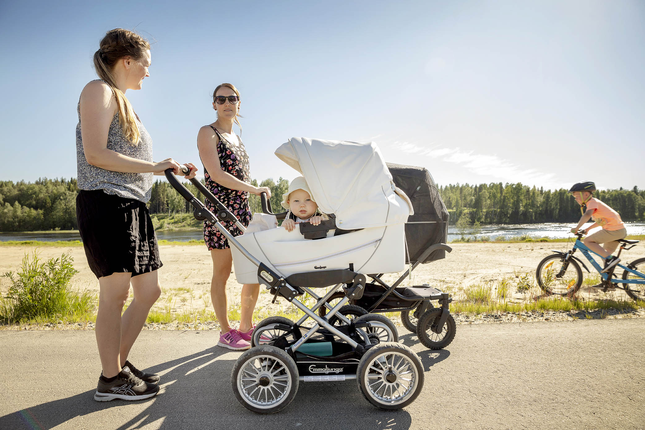 Två mammor på promenad på stranden med barnvagnar och en pojke på cykel. Foto.