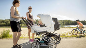 Två mammor på promenad på stranden med barnvagnar och en pojke på cykel. Foto.