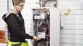 Kvinna med kort hår står vid värmecentral, en vit låda med många kablar