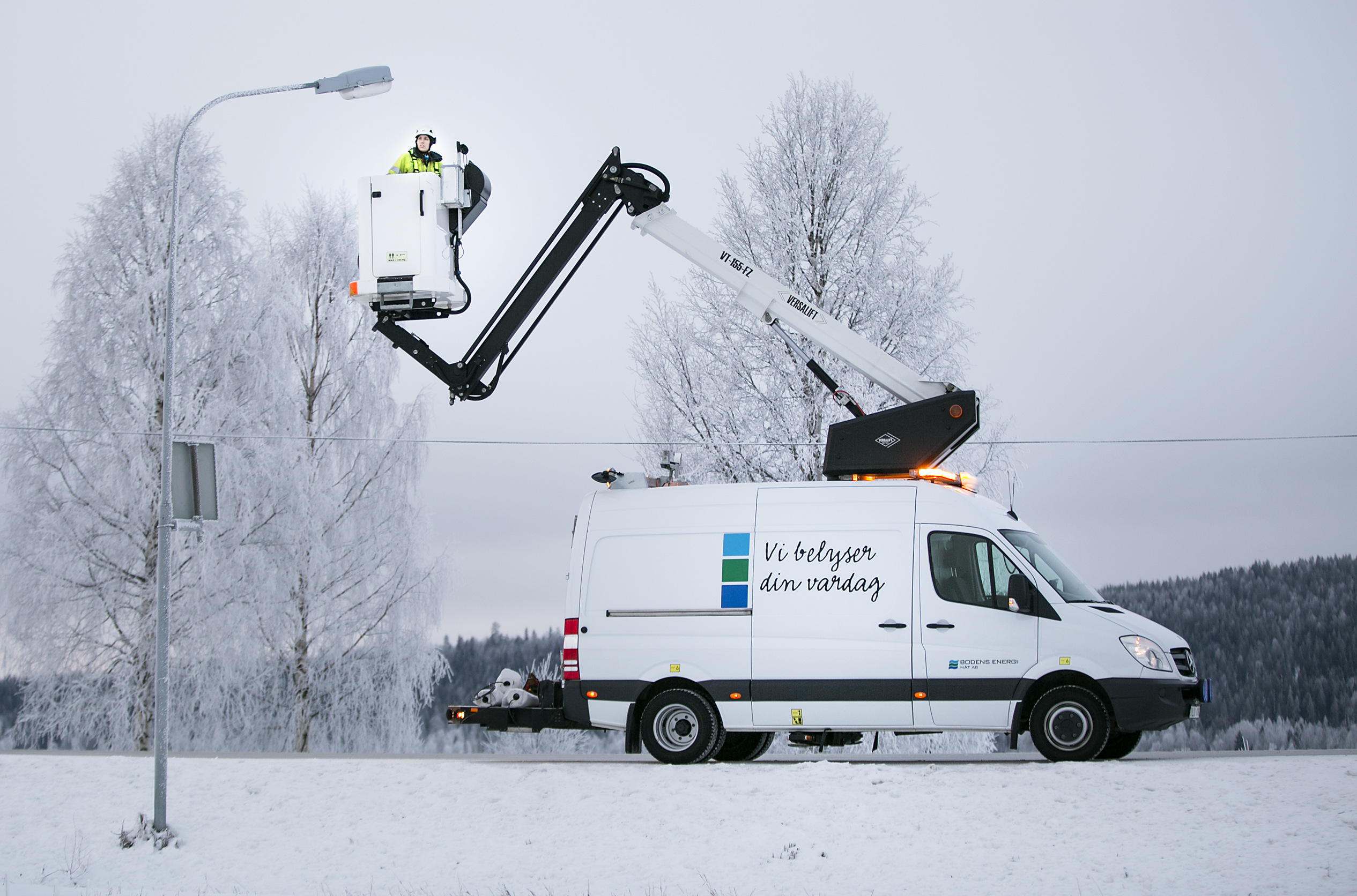 En tjej i arbetskläder och vit hjälp står i en skylift vid en gatubelysningsstolpe. Foto